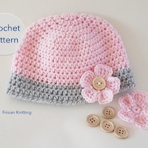 Op de afbeelding: Roze en grijze gehaakte baby muts met een bloemversiering en knopen. Haakpatroon.