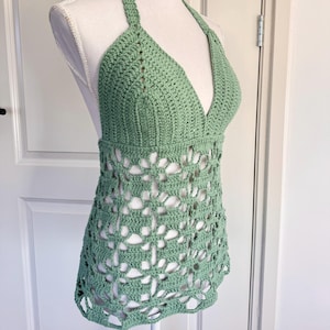 Op de afbeelding: Een saliegroene gehaakte haltertop met een diepe V-hals en openwerk ontwerp. De top wordt getoond op een mannequin, waarbij het ingewikkelde stikwerk en de vorm van het kledingstuk worden getoond.
