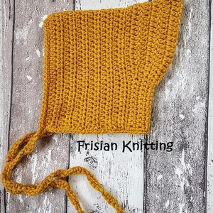 Op de afbeelding: Een mosterdgele gehaakte baby muts met twee lange banden. De muts is gemaakt met een eenvoudig steekpatroon en heeft een puntige vorm. De tekst "Frisian Knitting" is zichtbaar in de afbeelding.