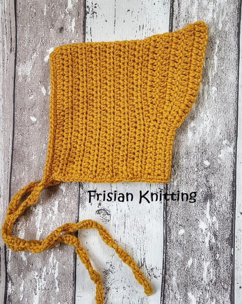 Op de afbeelding: Een mosterdgele gehaakte baby muts met twee lange banden. De muts is gemaakt met een eenvoudig steekpatroon en heeft een puntige vorm. De tekst "Frisian Knitting" is zichtbaar in de afbeelding.