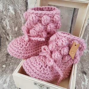 May include: A pair of pink crocheted baby booties with a textured design and a small brown leather tag with a heart on it. The booties are in a white wooden box.