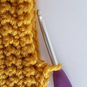 Op de afbeelding: Een close-up van een gehaakt stuk in mosterdgele kleur met een paarse haaknaald. De haaknaald is in het gehaakte stuk gestoken.
