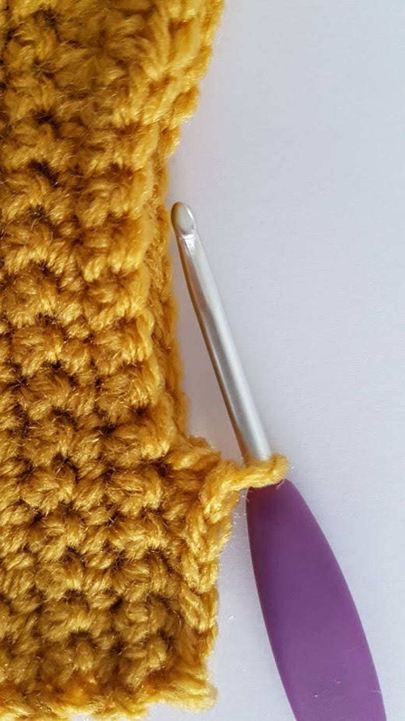 Op de afbeelding: Een close-up van een gehaakt stuk in mosterdgele kleur met een paarse haaknaald. De haaknaald is in het gehaakte stuk gestoken.