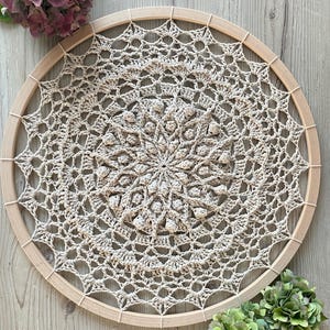 May include: A beige crocheted mandala with a wooden hoop, hanging on a light wood background. The mandala has a detailed design with a central circle of raised stitches.