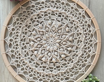 May include: A beige crocheted mandala with a wooden hoop, hanging on a light wood background. The mandala has a detailed design with a central circle of raised stitches.