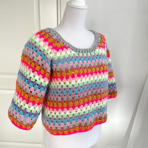 Peut inclure: Un haut en crochet coloré avec un motif rayé dans les tons de rose, violet, vert, bleu et marron. Le haut a un col rond et des manches courtes.