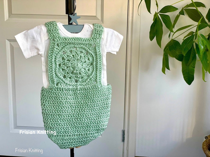 May include: A light green crocheted baby romper with a square-shaped design featuring a circular pattern in the center. The romper is on a white background.
