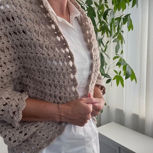 Peut inclure: Un châle en crochet beige avec un motif ajouré délicat. Le châle est porté sur une chemise blanche.