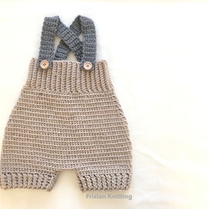 Op de afbeelding: Handgemaakte gehaakte baby-overall in een neutrale beige kleur met grijze bandjes. De overall heeft houten knopen en een geribbelde textuur. Het ontwerp omvat gekruiste bandjes en is geschikt voor een pasgeborene.