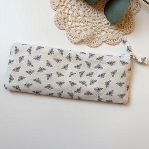 May include: A rectangular pencil case with a white background and a pattern of bees. The case has a white zipper and a small fabric loop. The bees are in a dark gray color.
