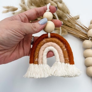 May include: A handmade macrame rainbow wall hanging with a brown, orange, and cream color scheme. The rainbow is accented with a wooden bead garland.