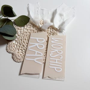 May include: Two rectangular beige acrylic bookmarks with the words "PRAY" and "WORSHIP" in white. The bookmarks are on a white surface with a beige lace doily, faux greenery, and a white fabric ribbon.