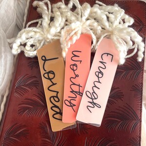 Puede incluir: Tres marcap&aacute;ginas con las palabras "Loved", "Worthy" y "Enough" escritas en letra negra sobre un diario de cuero marr&oacute;n con un patr&oacute;n de hojas de palma.