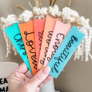 Puede incluir: Seis marcadores de libros de acr&iacute;lico de colores con diferentes nombres escritos en letra cursiva negra. Los marcadores est&aacute;n dispuestos en forma de abanico, con el marcador turquesa en la parte inferior y el marcador rosa en la parte superior.