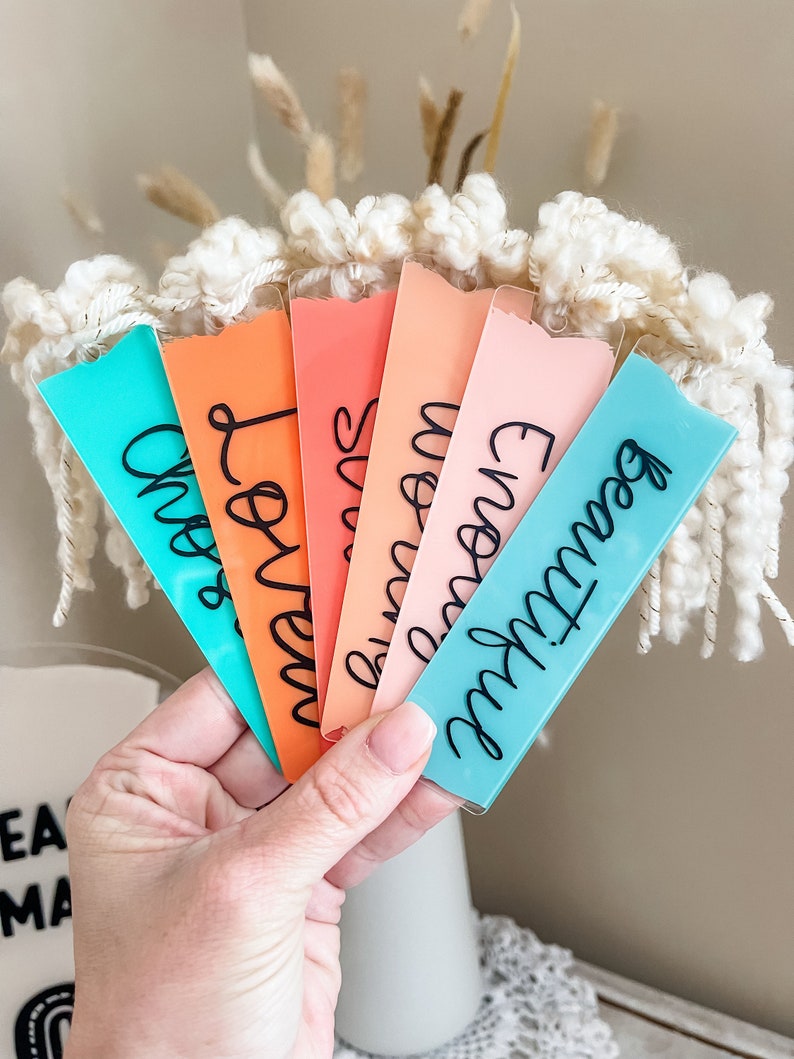 Puede incluir: Seis marcadores de libros de acr&iacute;lico de colores con diferentes nombres escritos en letra cursiva negra. Los marcadores est&aacute;n dispuestos en forma de abanico, con el marcador turquesa en la parte inferior y el marcador rosa en la parte superior.