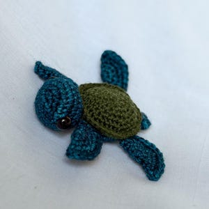 May include: A handmade crochet turtle in shades of teal and olive green. The turtle has a rounded head, a textured shell, and four flipper-like appendages. The eyes are small, dark beads. The turtle is lying on a white surface.