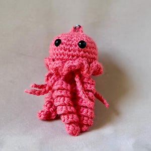 May include: A handmade, pink crocheted jellyfish with black safety eyes. The amigurumi jellyfish has a rounded head, ruffled edges, and dangling tentacles. This soft toy is a unique, handcrafted item.