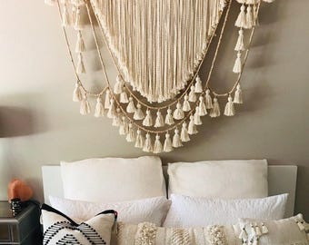 May include: A white macrame wall hanging with a triangular shape and multiple rows of tassels. The hanging is made with natural fibres and has a bohemian style.
