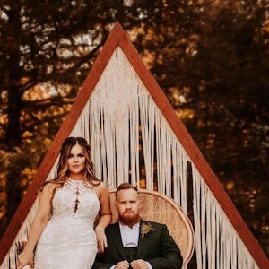 Op de afbeelding: Een bruid en bruidegom poseren voor een trouwfoto voor een houten driehoekige boog versierd met macramé. De bruid draagt een witte kanten trouwjurk en de bruidegom draagt een donkergroen pak. Ze staan op een terras van bakstenen.