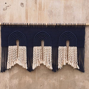 May include: A black and white macrame wall hanging with three arches. The arches are made of thick black cord and the bottom of the hanging has a fringe of white cord.
