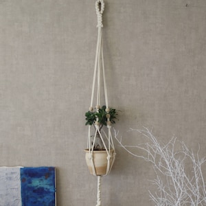 Könnte beinhalten: Ein Makramee-Pflanzenhänger mit einem weißen Quasten hängt an der Wand. Eine Topfpflanze mit grünen Blättern befindet sich im Hänger. Eine kleine Topfpflanze steht auf einem Holztisch mit schwarzen Metallbeinen. Ein weißer Zweig-Arrangement befindet sich im Hintergrund.