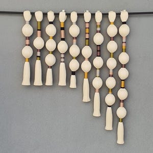 May include: A decorative wall hanging featuring a series of off-white, spherical beads and tassels suspended from a black rod. The beads are interspersed with colorful accents of pink, yellow, and brown. The background is a solid gray.