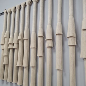 K&ouml;nnte beinhalten: Ein Wandbehang aus Naturfaserseil mit einem mehrstufigen Fransenmuster. Das Seil ist hellbeige und die Fransen sind in einem kaskadierenden Muster angeordnet.