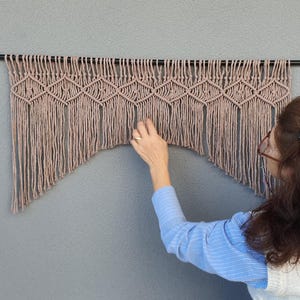 Könnte beinhalten: Beige-brauner Makramee-Wandbehang mit geometrischem Rautenmuster und Fransen, aufgehängt an einer schwarzen Metallstange. Die Wand hinter dem Makramee ist einfarbig grau. Das Textil ist etwa 100 cm breit.