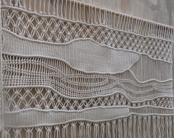 May include: A large, off-white macrame wall hanging with a woven pattern and fringe. The hanging is made of natural fibres and has a bohemian style.