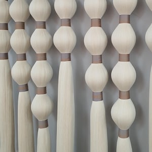 Puede incluir: Cortina de macramé hecha a mano, de color crema, con un patrón repetitivo de grandes nudos redondeados y largas borlas. La cortina está suspendida de una barra negra, creando un elemento decorativo texturizado y de tonos neutros para espacios interiores.