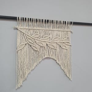 May include: An off-white macrame wall hanging featuring a central diagonal branch motif with leaf patterns. The piece is constructed from knotted cotton cord and is suspended from a thin, horizontal black metal rod, creating a pointed bottom edge.