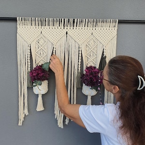 May include: Macrame wall hanging with three cream-colored plant holders, each holding a cluster of deep purple flowers. The macrame design features intricate knotting and fringe, creating a bohemian aesthetic. The wall hanging is mounted on a black rod.