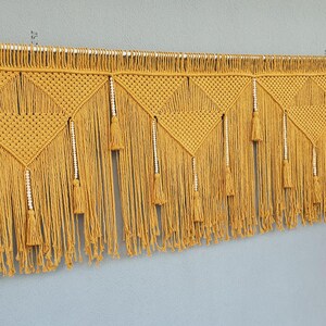 Può includere: Applique da parete in macramè giallo senape con motivi geometrici, nappe e dettagli di perline. L'opera d'arte tessile artigianale è sospesa a un'asta di metallo, aggiungendo un tocco bohémien all'arredamento.