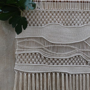 K&ouml;nnte beinhalten: Ein wei&szlig;er Makramee-Wandbehang mit geometrischem Muster und Fransen. Der Behang ist aus nat&uuml;rlichem Baumwollgarn gefertigt und ein beliebtes Dekoelement im Boho-Stil.