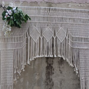 Op de afbeelding: Een crèmekleurige macramé wandhanger met ruitpatronen en franjes. Het handgemaakte textiel is versierd met een boeket witte en groene bloemen, wat een bohemien esthetiek creëert. De wanddecoratie wordt tentoongesteld tegen een verweerde betonnen muur.