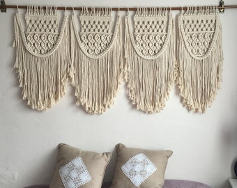 May include: A set of four macrame wall hangings in a cream colour, hanging from a wooden dowel rod. The hangings are made of knotted cords and have a fringe at the bottom. Two throw pillows with white lace designs are on a light purple bedspread.