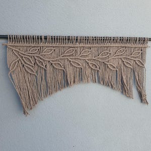 May include: A beige macrame wall hanging featuring a leaf design. The macrame is suspended from a dark metal rod. The leaf pattern is intricately knotted, with long fringe at the bottom. This handcrafted piece adds a touch of bohemian style to any space.