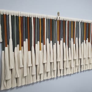 May include: A wall hanging made of yarn with tassels. The yarn is in shades of gray, green, orange, and white.