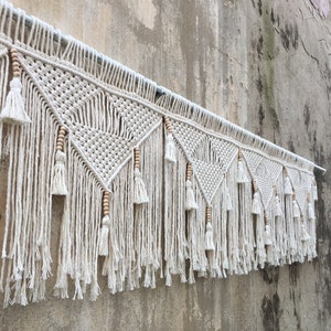 May include: A white macrame wall hanging with a geometric pattern and wooden beads. The hanging has a fringe of tassels at the bottom.