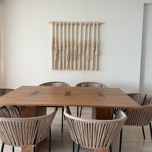K&ouml;nnte beinhalten: Ein Esstisch aus Holz mit einer nat&uuml;rlichen Holzoptik und einem gewebten Makramee-Wandbehang dar&uuml;ber. Der Tisch hat eine rechteckige Tischplatte und ist von sechs geflochtenen St&uuml;hlen mit schwarzen Metallbeinen umgeben.
