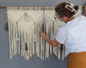 May include: Cream macrame wall hanging with a geometric pattern and fringe. The wall decor includes grey tassels and wooden beads. It's displayed against a grey wall, adding a bohemian touch. The piece would suit any room.