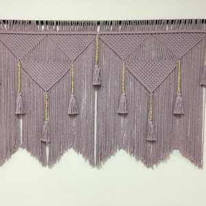 May include: A macrame wall hanging with a geometric pattern in a dusty rose colour. The hanging features wooden beads and tassels.