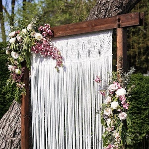 May include: A white macrame backdrop hangs from a wooden frame decorated with pink and white flowers. The backdrop is made of a series of long, thin cords that are knotted together to create a delicate and intricate pattern.