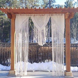 May include: A white macrame wedding arch hanging from a wooden pergola. The arch is made of knotted rope and hangs in front of a snowy background.