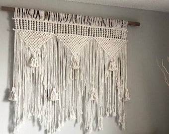 May include: A white macrame wall hanging with a geometric pattern and fringe. The hanging is made of cotton cord and has a wooden dowel at the top.
