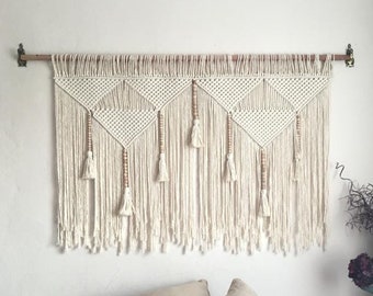 May include: A white macrame wall hanging with wooden beads and tassels. The hanging is made of cotton cord and is hung on a wooden dowel rod. The hanging is in front of a white wall and a bed with white and beige pillows.