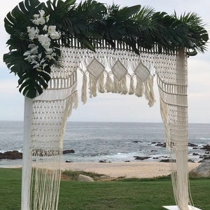 May include: A white macrame archway with a white wooden frame, decorated with green tropical leaves and white orchids. The archway is set up outdoors in front of a beach and ocean.