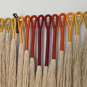 May include: A wall hanging made of natural fiber yarn with a gradient of colors from yellow to red. The yarn is tied to a wooden dowel rod and hangs in a cascade of strands.