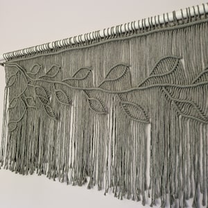 May include: A macrame wall hanging with a leafy design in a sage green color. The hanging is made with thick cotton cord and has a fringe at the bottom.