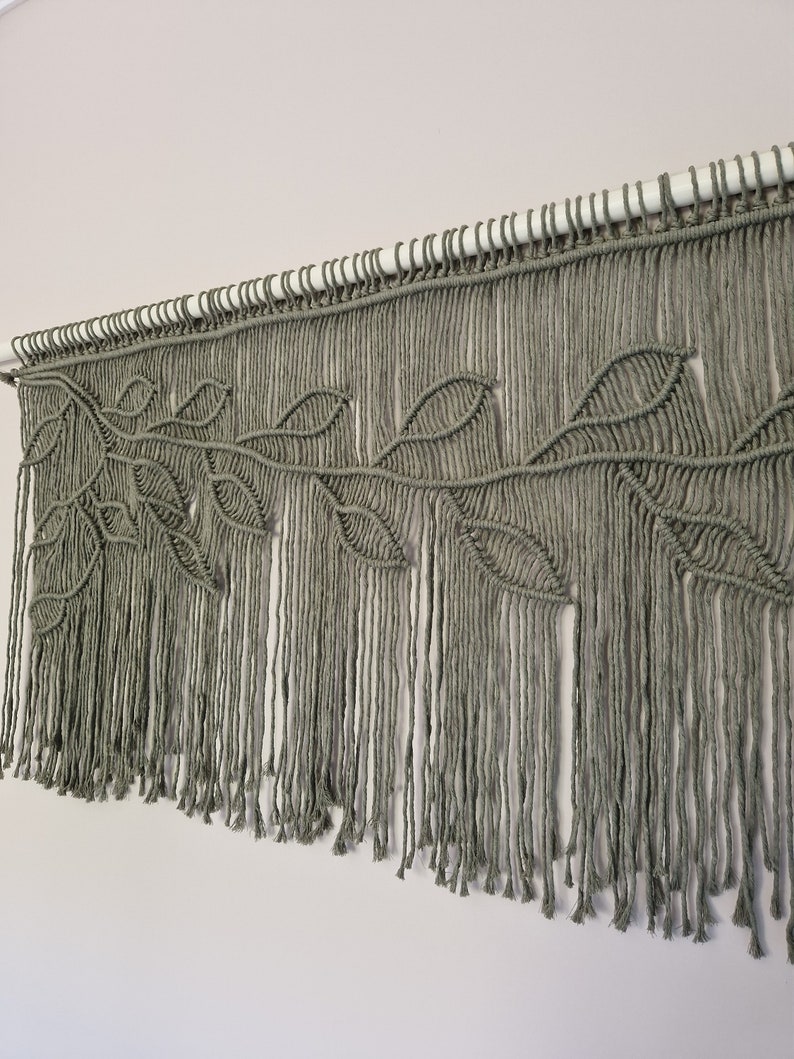 May include: A macrame wall hanging with a leafy design in a sage green color. The hanging is made with thick cotton cord and has a fringe at the bottom.